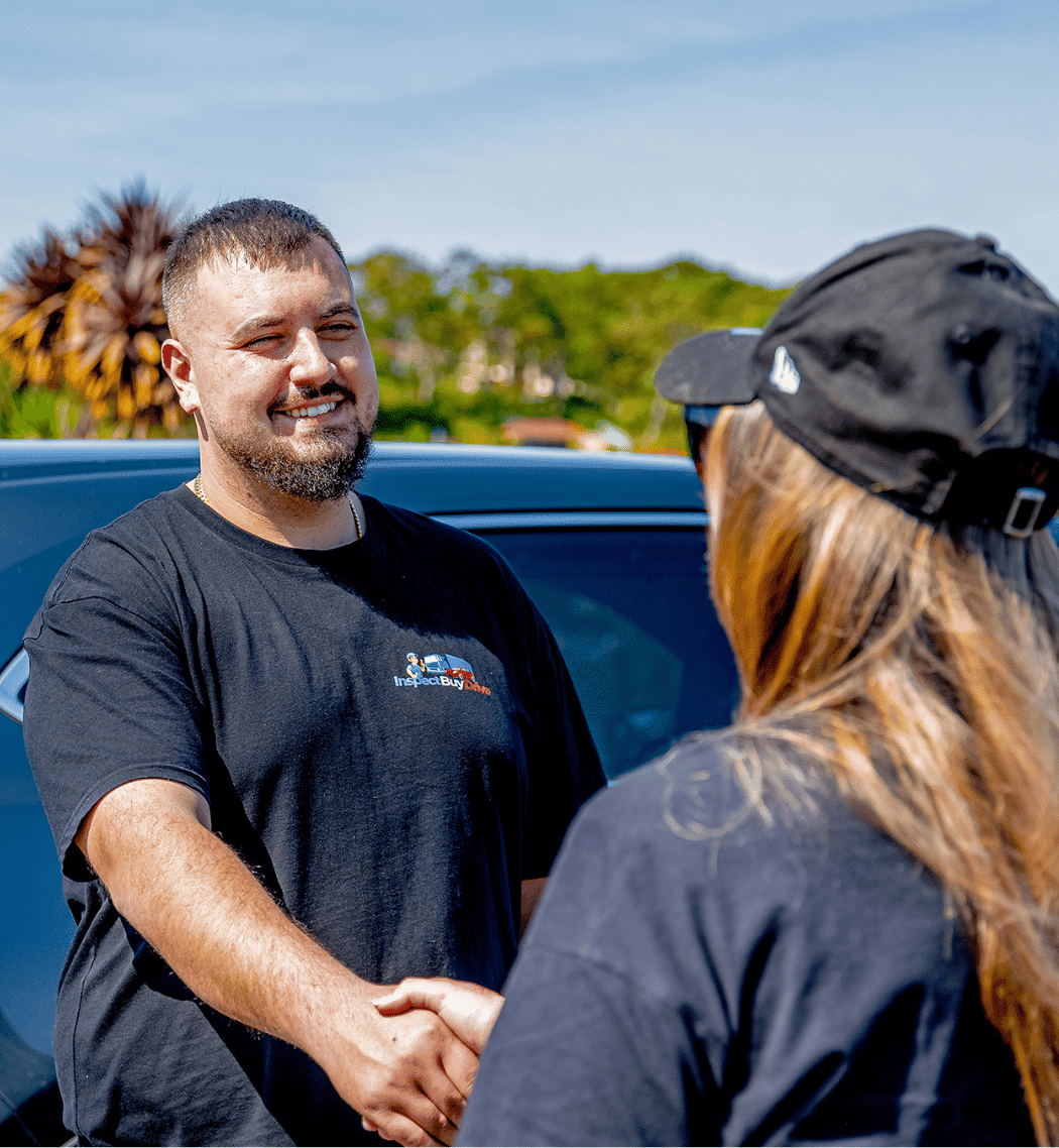 A smiling man in a black T-shirt shakes hands with a person in a black cap outdoors, beside a parked car and greenery under a clear blue sky.